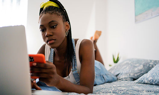 A teenage girl lies on her bed in front of her laptop with her mobile in her hand