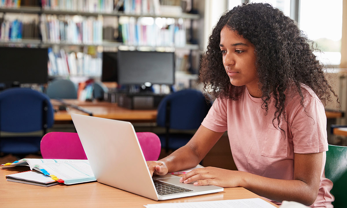 A teenage girl sits in a library working on her laptop