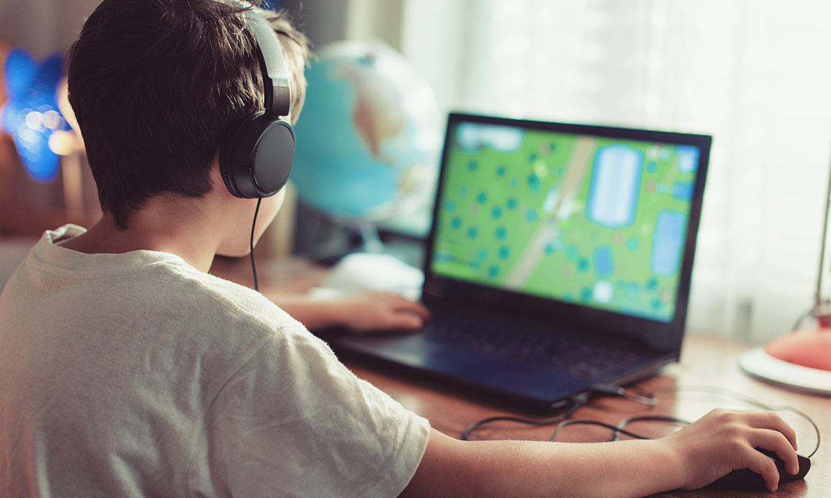 A boy sits at a desk on his laptop wearing headphones and playing a game 