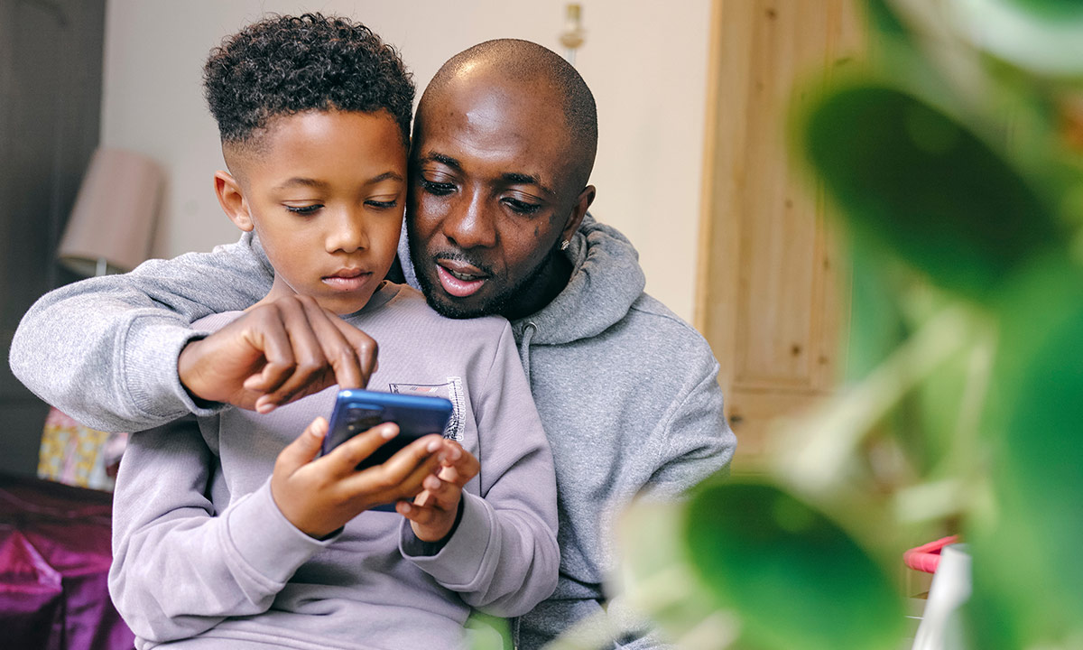 A father and son sit together at home playing on a phone