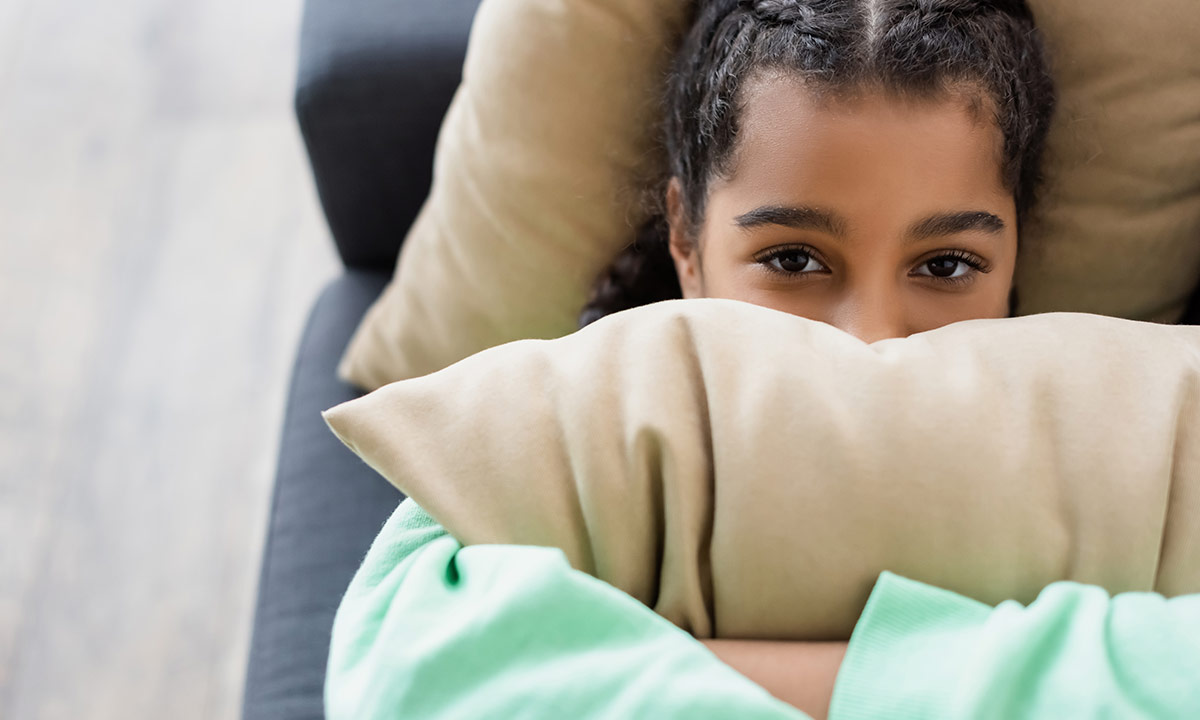 A teenage girl lies on the sofa hugging a pillow and looking directly at the camera